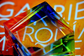 A transparent prism refracts light on an orange surface with white text partially visible through the prism, creating a vibrant display of colors including blue, green, yellow, and red.