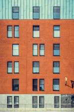 A modern building facade featuring a mix of architectural materials with a repeating pattern of windows. The top portion of the structure is covered with a bluish-green paneling, while the middle section showcases a red brick surface with uniformly aligned windows. The lower section has a grayish stone finish. An exterior lamp is mounted on the wall, casting a shadow.