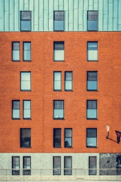 A modern building facade featuring a mix of architectural materials with a repeating pattern of windows. The top portion of the structure is covered with a bluish-green paneling, while the middle section showcases a red brick surface with uniformly aligned windows. The lower section has a grayish stone finish. An exterior lamp is mounted on the wall, casting a shadow.