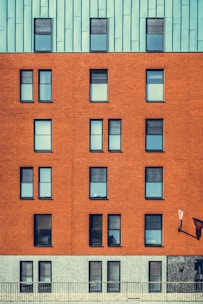 A modern building facade featuring a mix of architectural materials with a repeating pattern of windows. The top portion of the structure is covered with a bluish-green paneling, while the middle section showcases a red brick surface with uniformly aligned windows. The lower section has a grayish stone finish. An exterior lamp is mounted on the wall, casting a shadow.