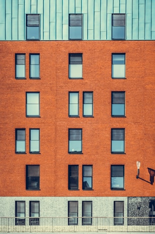 A modern building facade featuring a mix of architectural materials with a repeating pattern of windows. The top portion of the structure is covered with a bluish-green paneling, while the middle section showcases a red brick surface with uniformly aligned windows. The lower section has a grayish stone finish. An exterior lamp is mounted on the wall, casting a shadow.