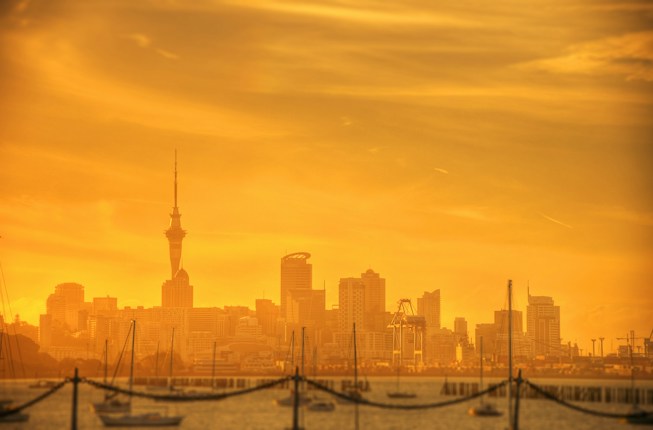 A city skyline is silhouetted against a warm, golden sky. The prominent needle-like tower stands out among other skyscrapers. In the foreground, silhouettes of boats and masts are visible across a body of water, creating a peaceful harbor scene.