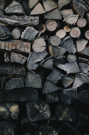 A stack of neatly arranged firewood featuring a variety of cut logs. The logs display different sizes and patterns of bark and wood grain, with most pieces split into manageable sections. The lighting is dim, highlighting the textures and natural tones of the wood.
