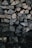 A stack of neatly arranged firewood featuring a variety of cut logs. The logs display different sizes and patterns of bark and wood grain, with most pieces split into manageable sections. The lighting is dim, highlighting the textures and natural tones of the wood.