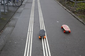 Two skateboards with orange wheels are situated on a paved path with double white lines. One board is black with a textured surface, and the other has a wooden finish with pink stripes. The path is bordered by a curb and greenery on one side, with parked bicycles visible nearby.