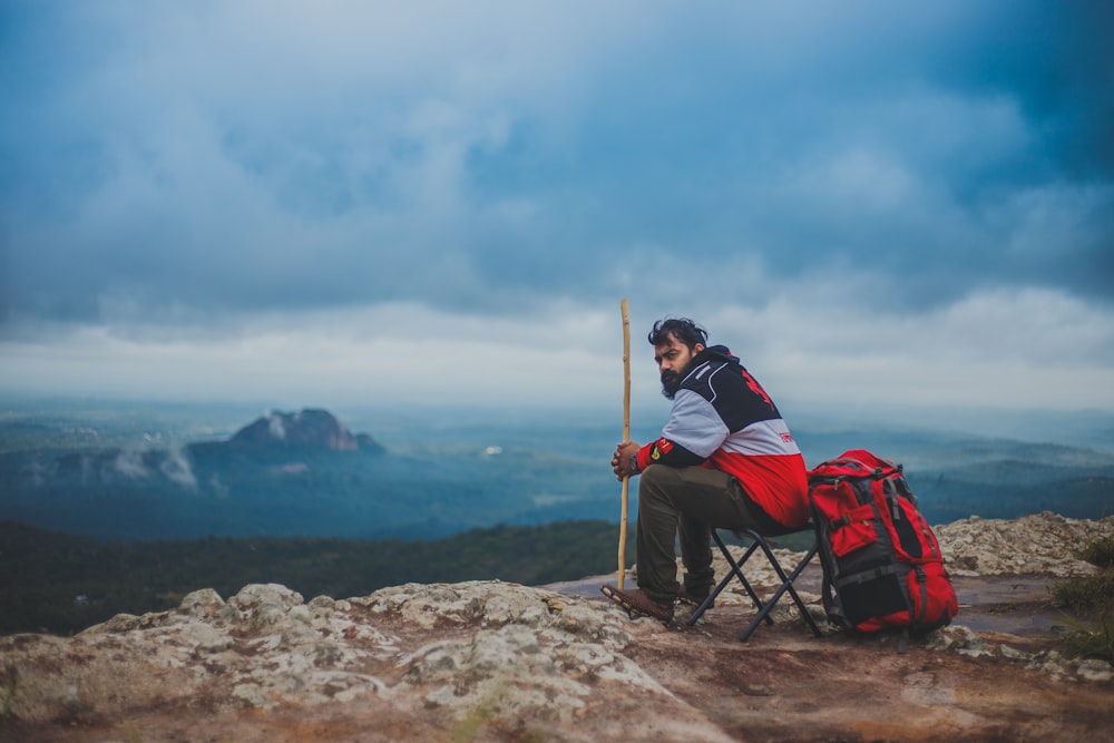 man sitting on camping chair while holding brown stick near mountain at daytime