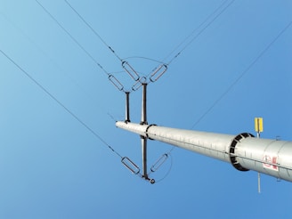 A tall metal power line pole extends upwards against a clear blue sky, with several electrical wires connected at the top.