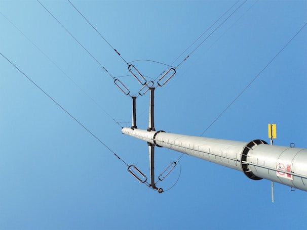 A tall metal power line pole extends upwards against a clear blue sky, with several electrical wires connected at the top.