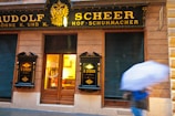 A storefront with elegant, classic black and gold signage displaying 'Rudolf Scheer Hof-Schuhmacher' and 'R. Scheer Söhne'. The entrance features wooden doors and illuminated windows, suggesting a warm, inviting atmosphere inside. A person holding a blue and white striped umbrella moves across the scene, indicating rainy weather.