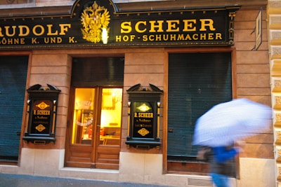 A storefront with elegant, classic black and gold signage displaying 'Rudolf Scheer Hof-Schuhmacher' and 'R. Scheer Söhne'. The entrance features wooden doors and illuminated windows, suggesting a warm, inviting atmosphere inside. A person holding a blue and white striped umbrella moves across the scene, indicating rainy weather.