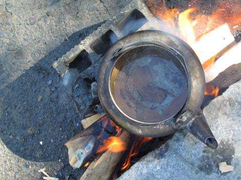 A metal kettle is placed on a makeshift stove made of bricks and wooden logs. The fire beneath the kettle emits bright orange flames and smoke can be seen rising from the heat. The surface around the setup is made of rough, dark concrete.
