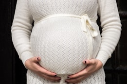 A person wearing a light-colored knit sweater with a ribbon tied around the waist cradles their pregnant belly with both hands. The background is dark, contrasting with the light-colored clothing.