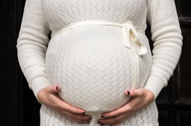 A person wearing a light-colored knit sweater with a ribbon tied around the waist cradles their pregnant belly with both hands. The background is dark, contrasting with the light-colored clothing.