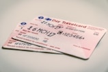 Two travel tickets are placed on a flat surface. The tickets are for a day pass, with printed text showing details like the date, type of travel, and zones. The predominant color of the tickets is a light pink, with black text and some blue and red symbols.