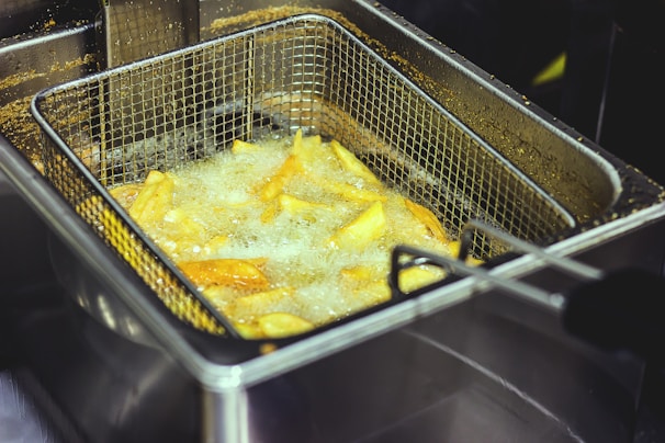 A metal basket filled with potato fries is submerged in hot oil inside a deep fryer. The fries are sizzling and creating bubbles due to the boiling oil. The background shows part of the fryer and some oil residue.