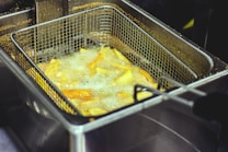 A metal basket filled with potato fries is submerged in hot oil inside a deep fryer. The fries are sizzling and creating bubbles due to the boiling oil. The background shows part of the fryer and some oil residue.