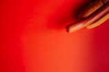 A hot dog protrudes from the right side against a vibrant red background. The hot dog is nestled inside a sliced bun, with a glossy sheen on the sausage.