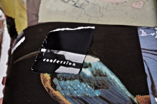 A torn black piece of paper with the word 'confession' typed on it is placed on top of a colorful, abstract background. The background includes shades of blue, brown, and other mixed colors, possibly part of a painting.