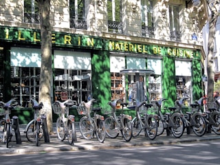 A row of rental bicycles parked in front of a cookware store with a green facade. The storefront window displays various kitchen appliances and cookware. Shadows cast by nearby trees create intricate patterns on the building wall and the bicycles.