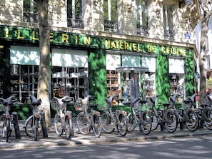 A row of rental bicycles parked in front of a cookware store with a green facade. The storefront window displays various kitchen appliances and cookware. Shadows cast by nearby trees create intricate patterns on the building wall and the bicycles.