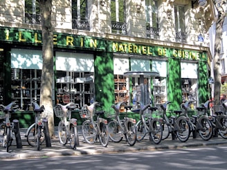 A row of rental bicycles parked in front of a cookware store with a green facade. The storefront window displays various kitchen appliances and cookware. Shadows cast by nearby trees create intricate patterns on the building wall and the bicycles.