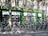 A row of rental bicycles parked in front of a cookware store with a green facade. The storefront window displays various kitchen appliances and cookware. Shadows cast by nearby trees create intricate patterns on the building wall and the bicycles.