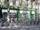 A row of rental bicycles parked in front of a cookware store with a green facade. The storefront window displays various kitchen appliances and cookware. Shadows cast by nearby trees create intricate patterns on the building wall and the bicycles.