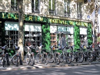 A row of rental bicycles parked in front of a cookware store with a green facade. The storefront window displays various kitchen appliances and cookware. Shadows cast by nearby trees create intricate patterns on the building wall and the bicycles.