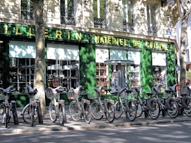A row of rental bicycles parked in front of a cookware store with a green facade. The storefront window displays various kitchen appliances and cookware. Shadows cast by nearby trees create intricate patterns on the building wall and the bicycles.
