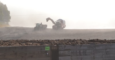 In a foggy or dusty agricultural setting, a tractor and a combine harvester operate in a field with large crates filled with harvested crops in the foreground. The atmosphere is hazy, suggesting early morning or evening work.