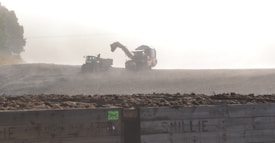 In a foggy or dusty agricultural setting, a tractor and a combine harvester operate in a field with large crates filled with harvested crops in the foreground. The atmosphere is hazy, suggesting early morning or evening work.