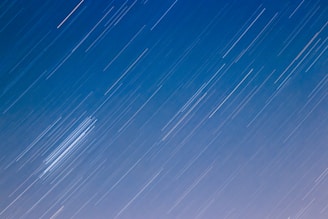 A night sky with long, streaking star trails from a long-exposure photograph against a gradient from blue to light purple.