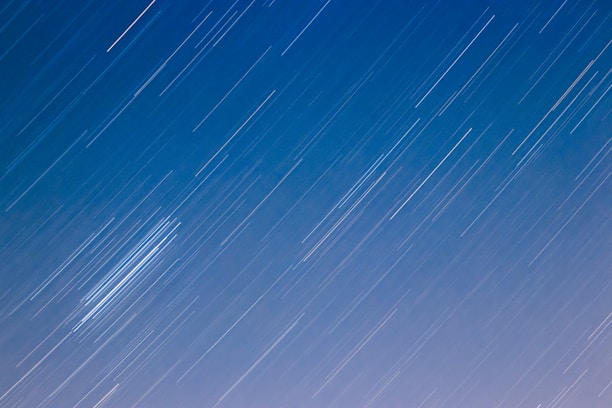 A night sky with long, streaking star trails from a long-exposure photograph against a gradient from blue to light purple.