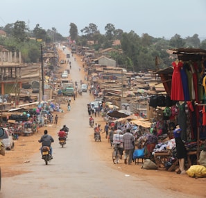 A bustling street market with numerous stalls displaying various goods, including clothing and accessories. People are walking, biking, and riding motorcycles along the dirt road. The area is lively with a mix of market activity and traffic, while the surroundings have a semi-rural appearance with greenery in the background.