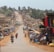 A bustling street market with numerous stalls displaying various goods, including clothing and accessories. People are walking, biking, and riding motorcycles along the dirt road. The area is lively with a mix of market activity and traffic, while the surroundings have a semi-rural appearance with greenery in the background.
