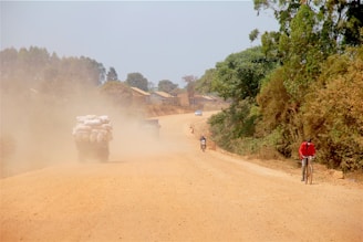A dusty rural road stretches into the distance with a truck carrying large sacks, stirring up a cloud of dust. On the right, a person in a red jacket rides a bicycle along the edge of the road, while two motorcyclists can be seen further down. Trees line the road, providing greenery amidst the dry terrain.