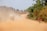 A dusty rural road stretches into the distance with a truck carrying large sacks, stirring up a cloud of dust. On the right, a person in a red jacket rides a bicycle along the edge of the road, while two motorcyclists can be seen further down. Trees line the road, providing greenery amidst the dry terrain.