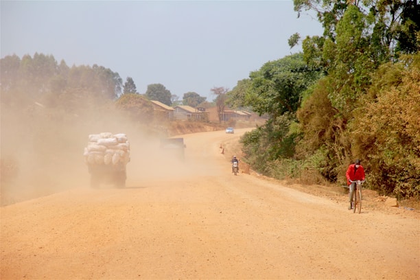 A dusty rural road stretches into the distance with a truck carrying large sacks, stirring up a cloud of dust. On the right, a person in a red jacket rides a bicycle along the edge of the road, while two motorcyclists can be seen further down. Trees line the road, providing greenery amidst the dry terrain.
