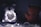A dimly lit scene featuring two teddy bears facing each other. The bear on the right appears to be reddish-brown and is visible from the back, while the other is obscured by soft focus and holds a heart-shaped string of white lights.