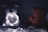 A dimly lit scene featuring two teddy bears facing each other. The bear on the right appears to be reddish-brown and is visible from the back, while the other is obscured by soft focus and holds a heart-shaped string of white lights.