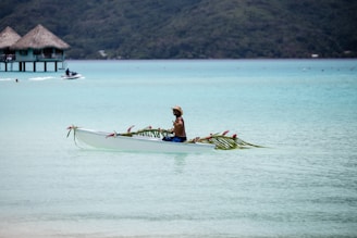A person paddles a decorated canoe in a serene, turquoise lagoon with thatched overwater bungalows in the background. The canoe is adorned with palm leaves and pink flowers, and the person appears relaxed, wearing a hat and swim trunks.