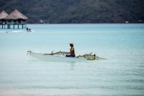 A person paddles a decorated canoe in a serene, turquoise lagoon with thatched overwater bungalows in the background. The canoe is adorned with palm leaves and pink flowers, and the person appears relaxed, wearing a hat and swim trunks.