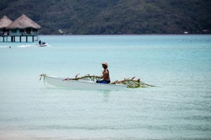 A person paddles a decorated canoe in a serene, turquoise lagoon with thatched overwater bungalows in the background. The canoe is adorned with palm leaves and pink flowers, and the person appears relaxed, wearing a hat and swim trunks.