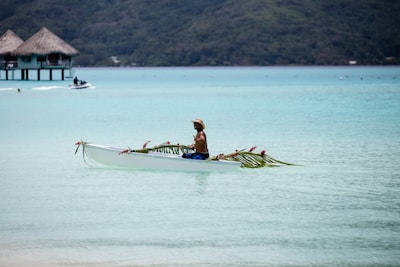 A person paddles a decorated canoe in a serene, turquoise lagoon with thatched overwater bungalows in the background. The canoe is adorned with palm leaves and pink flowers, and the person appears relaxed, wearing a hat and swim trunks.