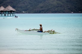 A person paddles a decorated canoe in a serene, turquoise lagoon with thatched overwater bungalows in the background. The canoe is adorned with palm leaves and pink flowers, and the person appears relaxed, wearing a hat and swim trunks.