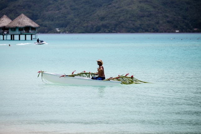 A person paddles a decorated canoe in a serene, turquoise lagoon with thatched overwater bungalows in the background. The canoe is adorned with palm leaves and pink flowers, and the person appears relaxed, wearing a hat and swim trunks.