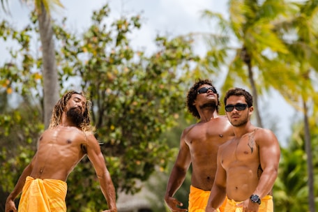 Three men are standing shirtless outdoors, wearing bright orange shorts. They are surrounded by tropical vegetation, including palm trees and other greenery. Each man displays a confident posture, with one of them wearing sunglasses.