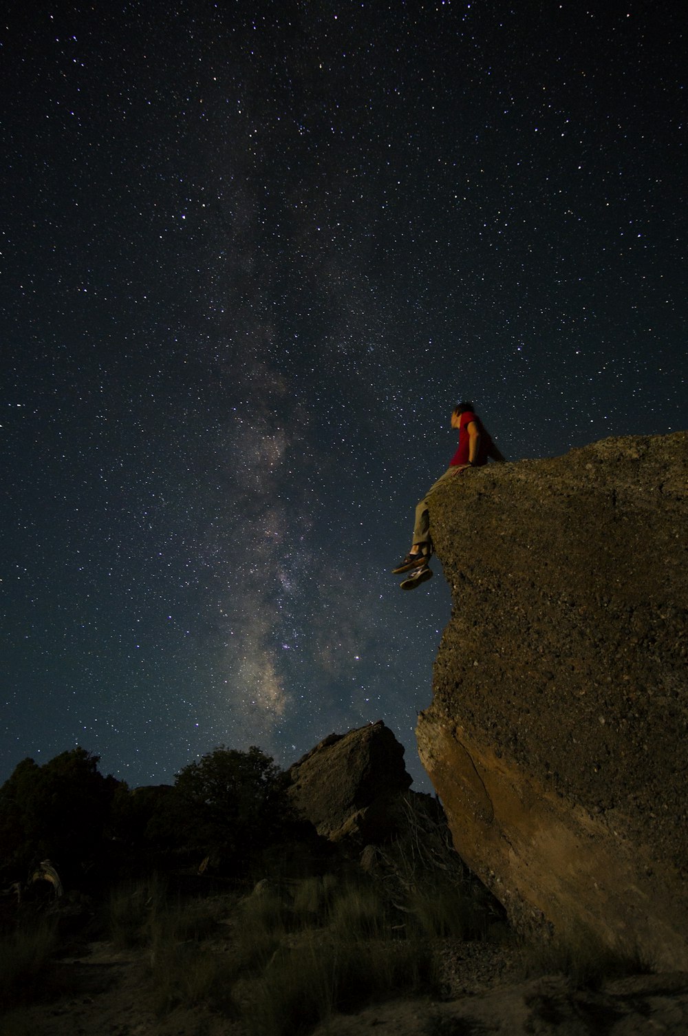 person sitting on edge of large rock under starry night sky