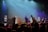 A lively band performing on stage with colorful lights and an engaged audience dancing.