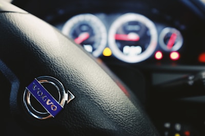 The close-up of a car steering wheel featuring the Volvo logo with dashboard dials and indicators blurred in the background.